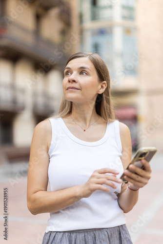 Free Download Brunette woman with smartphone walks along city street, spends time in motion, walks and enjoys sunny warm day. Woman wanders through city streets, sticks on phone, free time from work, leisure.. - GFXLoad