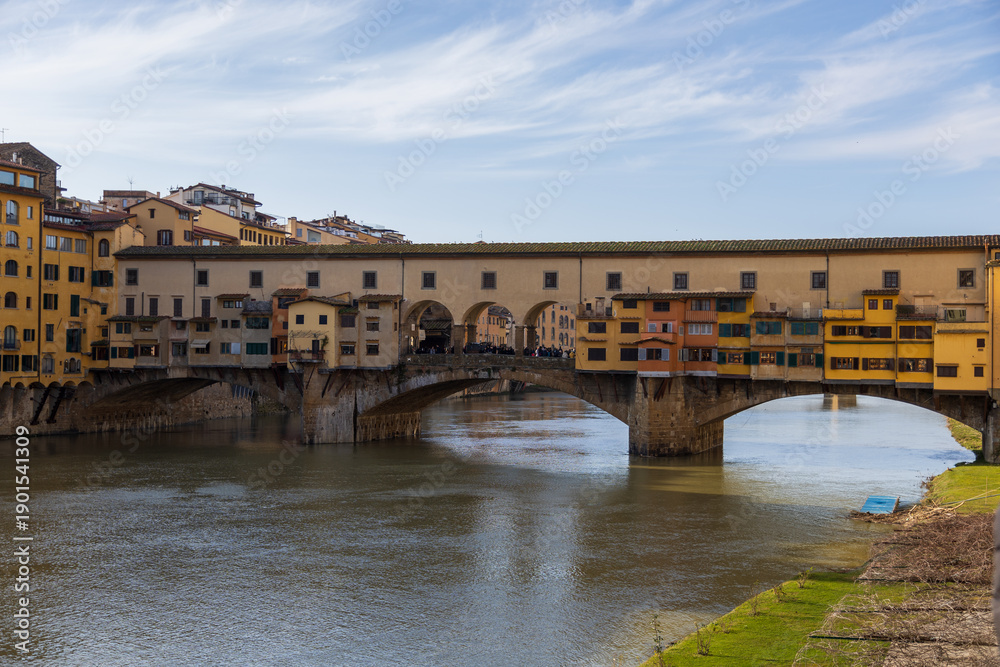 Naklejka premium Florence Bridge Scene. Colorful Riverside Structures With Reflection. Sunlit Cityscape Featuring Architecture And Water Reflections In Florence