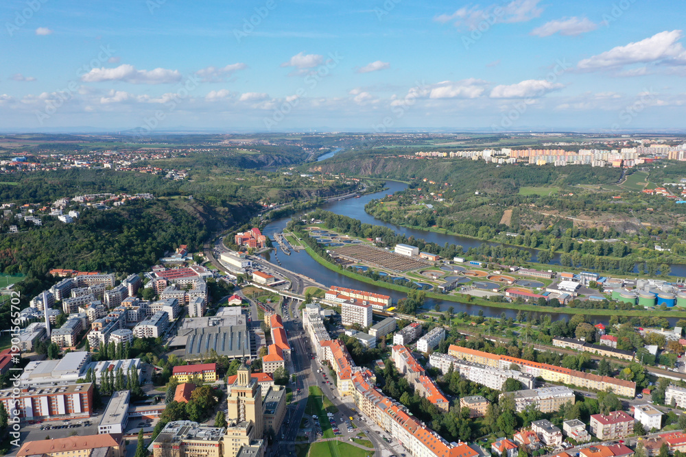 Fototapeta premium Aerial view of Prague with the Vltava River flowing through Dejvice, showing urban districts, green hills, and wastewater treatment plant under clear sky.