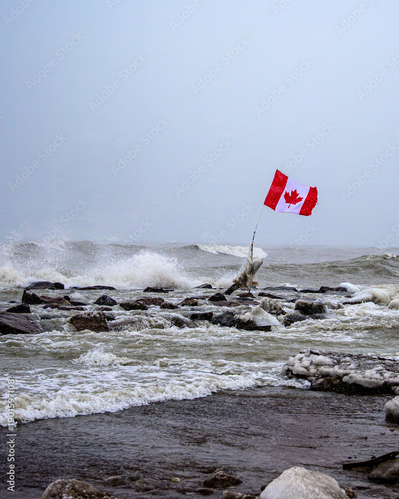 Obraz premium Vertical view of a Canadian flag fluttering in intense winter wind above a rough Lake Ontario.