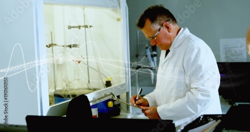 Writing senior chemist wearing white lab coat taking notes at bench with fume hood flask