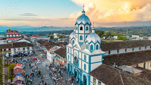 An aerial shot showcases a vibrant blue and white church with a crowd gathered below, set against a dramatic sunset sky.