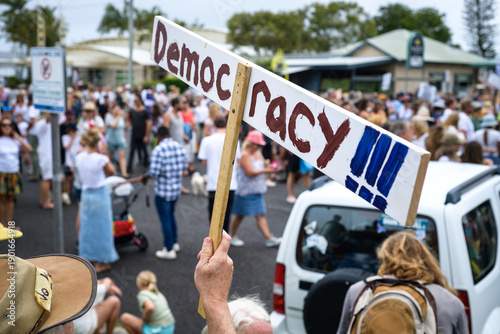 A person at an outdoor public rally holds a large wooden sign that reads 