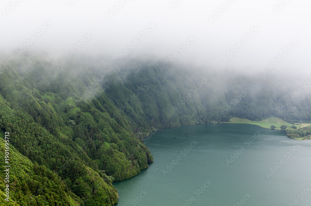Fototapeta premium Volcanic crater lake surrounded by dense fog and green forest in Sete Cidades, San Miguel, Azores. Misty atmosphere with caldera views and serene water.