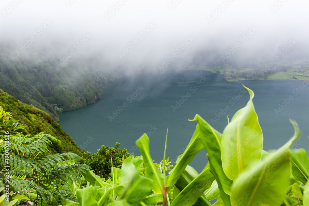 Naklejka premium Volcanic lake surrounded by lush greenery in Sete Cidades crater. Mist partially covers the landscape. View from elevated point in San Miguel, Azores.