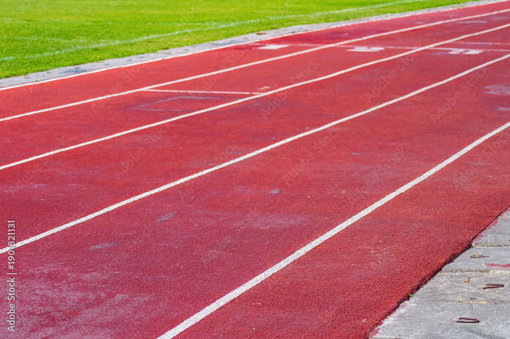 Fototapeta premium Red running track with lanes 3 to 5 marked in white, bordered by grass and concrete, ideal for race visuals.