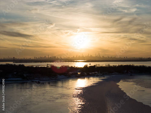 Distant manhattan skyline silhouetted against a vibrant sunset sky, reflecting golden light on the partially frozen manhasset bay and snowy shoreline in port washington, new york