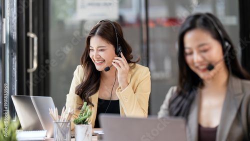 Konstfotografi Asian customer service woman smiling and wearing headset with colleague working in background at modern call center