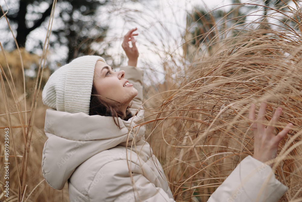 Fototapeta premium Woman in a winter coat and beanie among tall dried grass outdoors, enjoying the chilly air and nature in a peaceful winter landscape.
