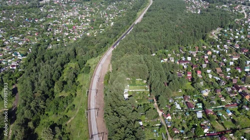 Electric commuter train vanishing into dense green forest along curving tracks, viewed from high above. Rural journey