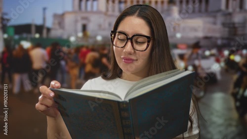 Woman with glasses reading a large hardcover book with a slight smile while standing on a busy street at dusk; learning focus discovery serenity.