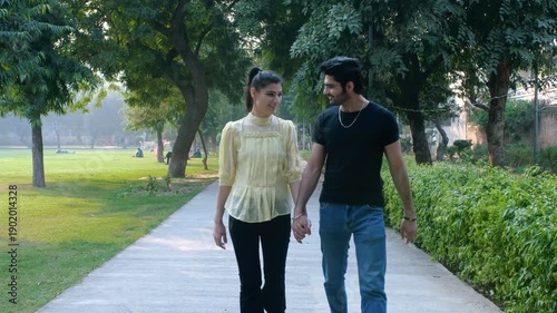 A romantic boyfriend- girlfriend hold hands while walking together on a sidewalk - pedestrians, pavement. A cheerful young romantic couple in the city park - a leisure time, pleasant weather, relation