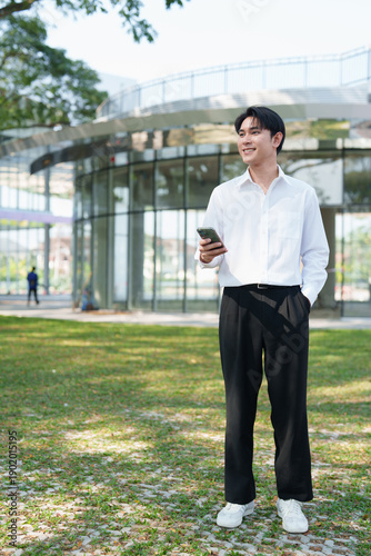 Fotografía Young man smiling standing outdoors, holding a smartphone, and looking away, wit