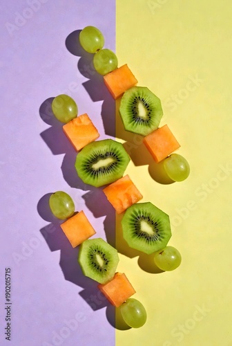 Vertical pattern of fresh fruit on wooden skewers, healthy kiwi and grape snacks on purple and yellow background, candid lifestyle food illustration
