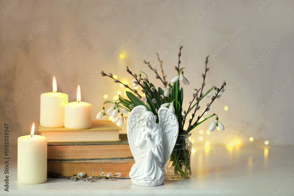 Naklejka premium praying angel figurine, burning candles, books, snowdrop flowers and willow in vase on table. Easter, Palm Sunday holidays concept. Christian church fest. symbol of faith in God, Lent, religion