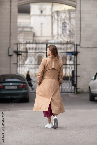Trenchcoat Woman Sneakers: Rear view of woman walking towards gate with church in background daytime.