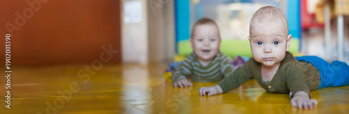 Two baby twin brothers are crawling on a bright yellow floor. One twin is closer to the camera while the other is in the background. The room has a playful environment. Empty space for text