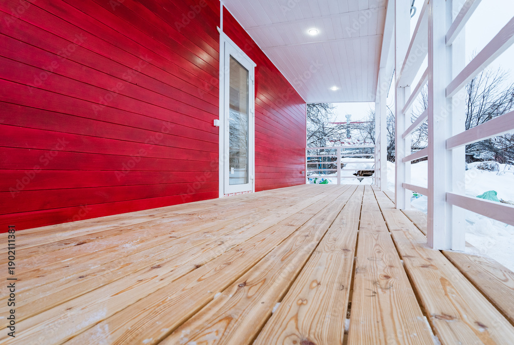 Fototapeta premium Wooden Porch With Red Exterior Wall And Pine Deck Overlooking Snowy Landscape, Open Railing And Bright Winter Light Create Inviting Seasonal Outdoor Space And Modern Cabin Vibe