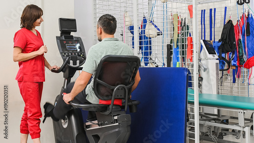 Obraz na płótnie A middle-aged man exercises on a stationary bike under the guidance of a physical therapist