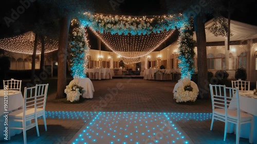 An elegant nighttime celebration illuminated by soft lights and floral arrangements. Tables are set with white linen and chairs. The scene includes a light path