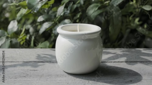 A white, ceramic candle sits on a weathered wooden surface, with a green leafy background. The candle has a single wick