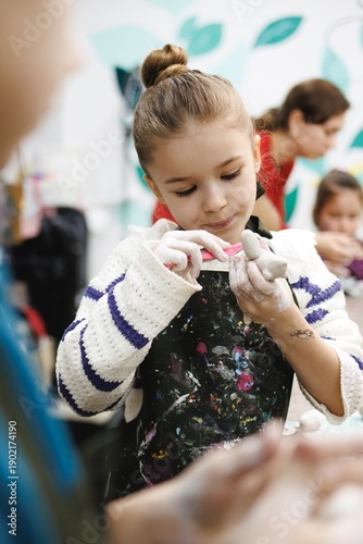 Young girl focused on shaping clay at a creative workshop with bright colors and other children around her
