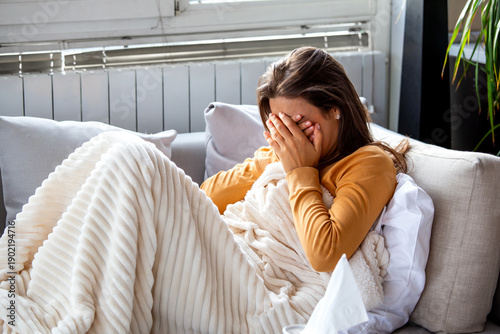 Sick woman crying on sofa at home. Sad female covering face with hands while wrapped in blanket feeling unwell and depressed. Healthcare and medicine concept with tissues and white soft blanket.