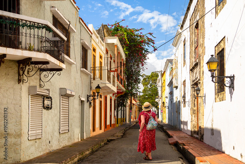 Photography A traveler in a red dress wanders down a sun-drenched, historic street in Santo Domingo, framed by colonial architecture and charming balconies