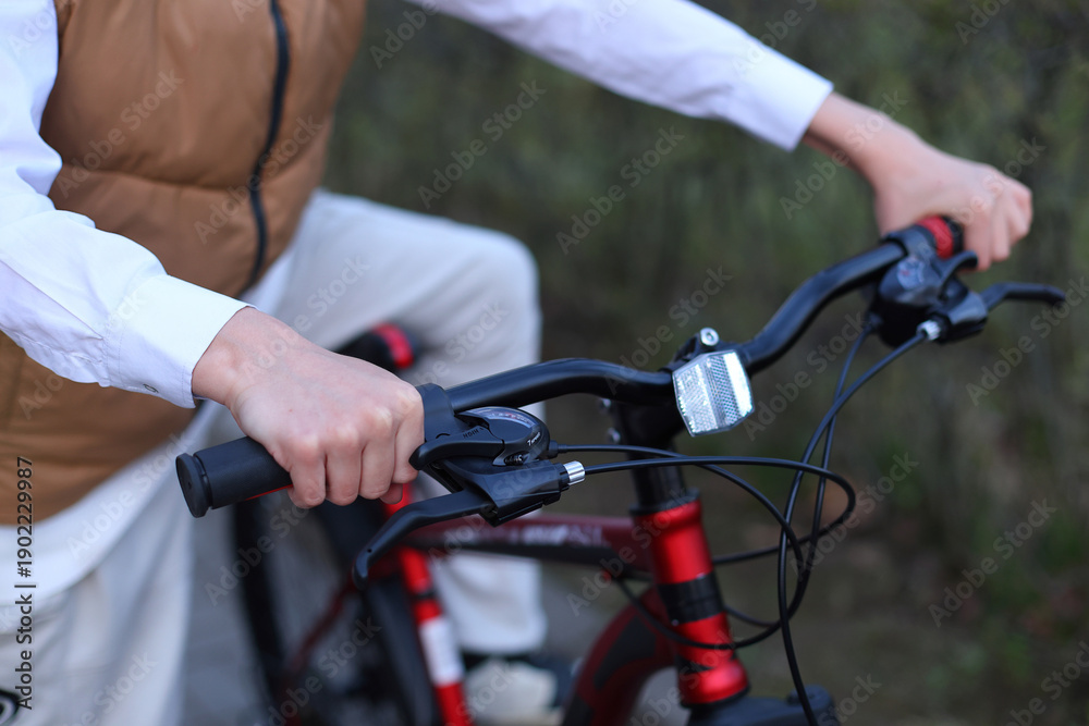 Obraz premium Close up of a boys hands gripping the handlebars of a red bike, brake levers, gear shifter, and reflector visible, poised for a ride in soft outdoor light.