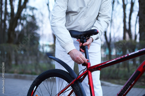 A person in a white jacket adjusts the bike saddle height on a red bicycle, focused, practical outdoor maintenance amid bare trees and soft daylight.