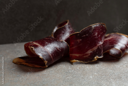 Elegant curls of Spanish cured beef cecina presented on a dark stone surface in a sophisticated low-light setting. Premium artisanal charcuterie with rich textures and deep tones, evoking luxury dinin