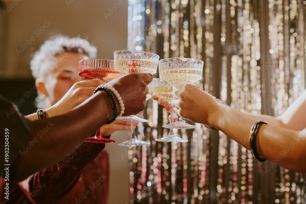 Fototapeta premium Diverse group of young adult and middle aged people raising champagne glasses in celebratory toast, hands and arms visible with various skin tones, festive background shimmering