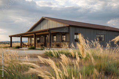 Beautiful Barndominium house stands in grassy field near mountains at sunset with clouds above and warm light shining down