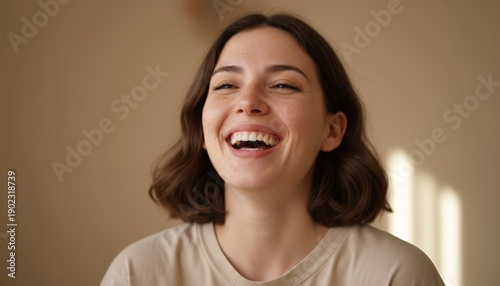 Young woman with freckles expressing happiness and positivity, laughing openly and sincerely, radiating a carefree and authentic emotion against a plain background with sunlight shadows