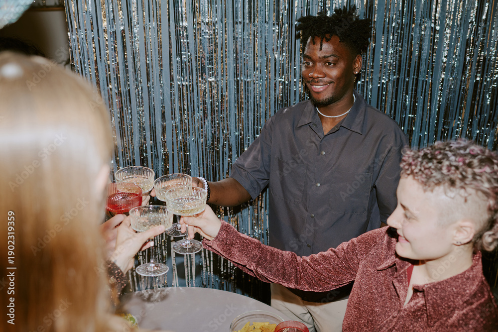 Fototapeta premium Group of young adults including Black man and Caucasian women clinking glasses and smiling during celebration, standing together in front of metallic streamers at festive event