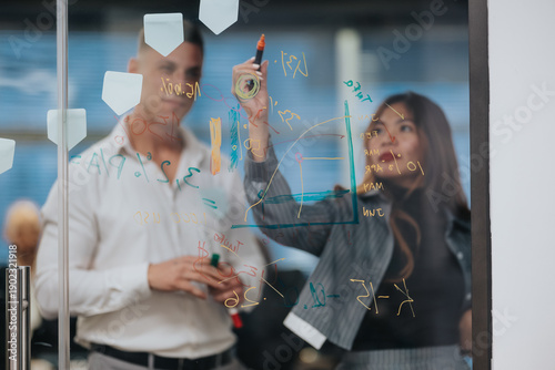 Canvas Print Two business workers write on a glass board with markers, drawing charts and notes