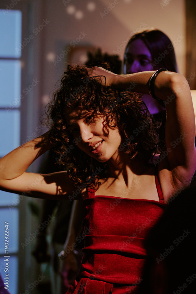Naklejka premium Caucasian young adult woman dancing with hands in curly hair at indoor party, smiling and enjoying music, other people visible in background, festive social gathering atmosphere