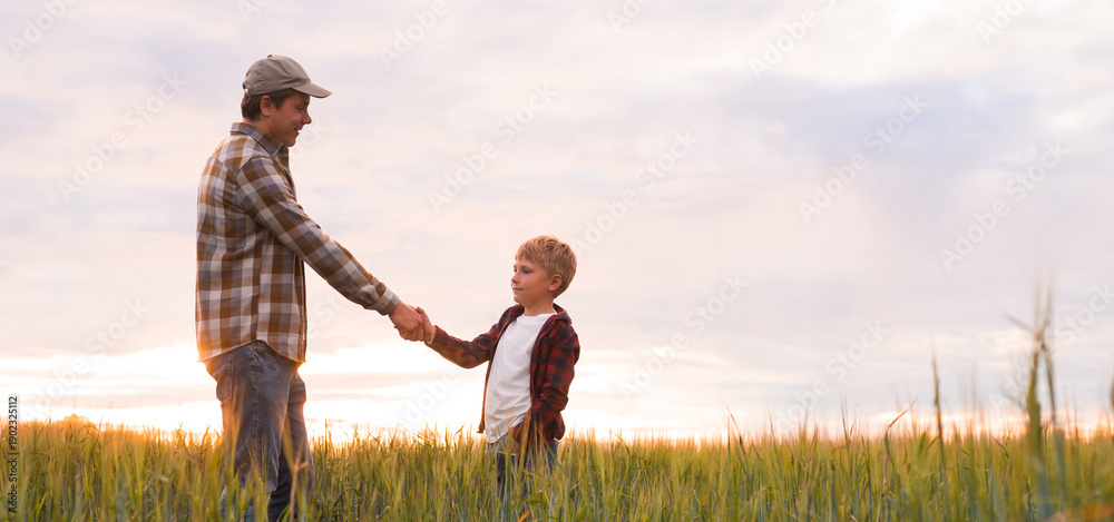 © Acronym - Farmer and his son in front of a sunset agricultural landscape. Man and a boy in a countryside field. Fatherhood, country life, farming and country lifestyle concept.