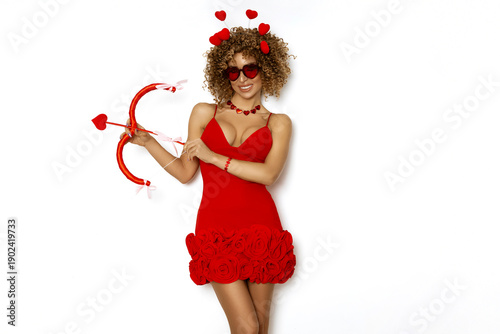 Playful young woman with curly hair wearing red heart sunglasses and red dress holding cupid's bow for valentine's day celebration on white background