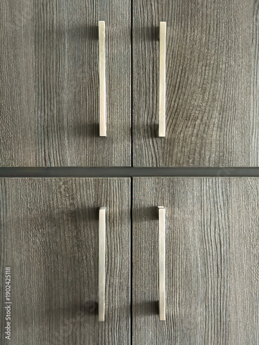 Set of four sleek nickel cabinet pulls on identical thermolaminated doors of contemporary kitchen cabinets in natural window lighting. Motifs of statement hardware and residential decor.