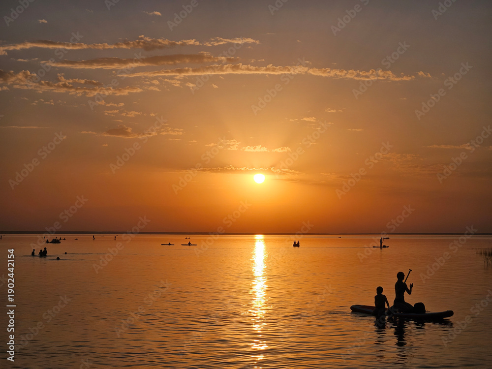Naklejka premium Group of people or Boy and a woman are paddling on a raft in the ocean. The sun is setting in the background