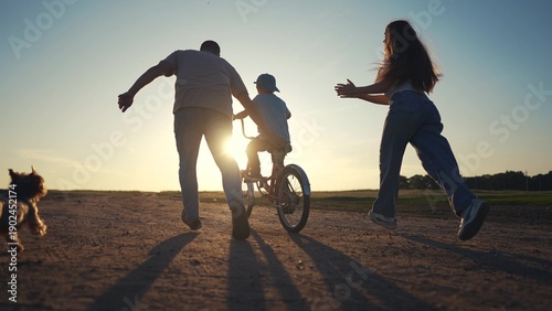 Family on dirt road at sunset. Family teaches their son how to ride bike. A father and child are riding a bicycle. A family on a gravel road during lifestyle sunset.