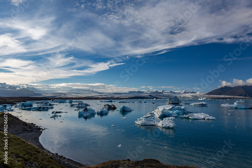 Blue and white icebergs drift across Jökulsárlón glacier lake as warm afternoon light settles over Southern Iceland’s sweeping glacier and rugged mountains rising quietly beyond the still lagoon