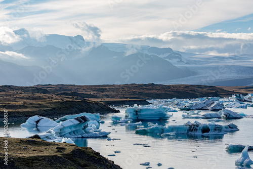Blue and white icebergs drift across Jökulsárlón glacier lake as soft evening light settles over Southern Iceland’s rugged hills and the distant glacier rising quietly beyond the mirrored water