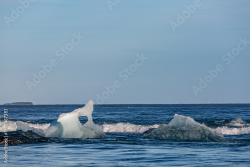 Shimmering ice fragments drift along the shore of Diamond Beach near Jökulsárlón glacier lake as soft afternoon light glows across Southern Iceland’s quiet waves and distant volcanic coastline