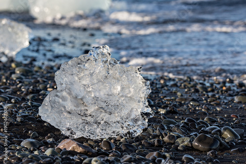 A crystal‑clear shard of ice rests on the dark pebbled shore of Jökulsárlón glacier lake as gentle sunrise light glows across Southern Iceland’s quiet waves and rugged coastline