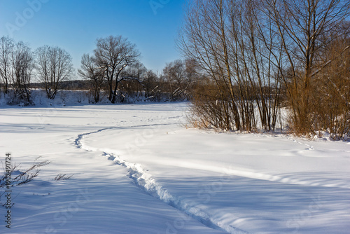 Canvastavla Footprints wind across a pristine snowscape towards a frozen riverbank under a b