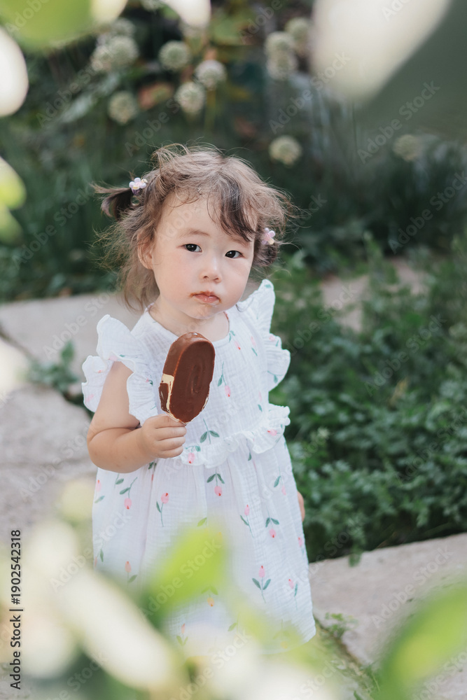 Naklejka premium Adoorable girl smiling and holding ice cream in hand in garden with flowers on background