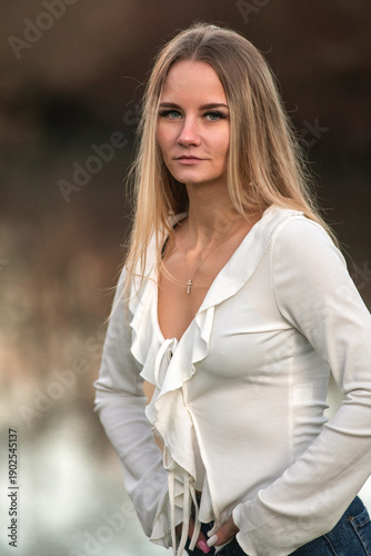 Young woman with long blonde hair wearing a white blouse and blue jeans stands by a reflective body of water in a natural outdoor setting