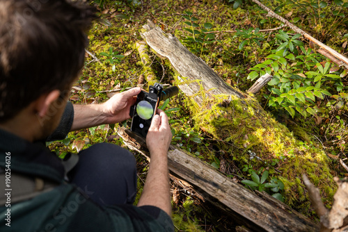 A first-person perspective (POV) over the shoulder of a man holding a smartphone with a macro lens attachment, displaying a sharp, magnified image of green moss on the screen.